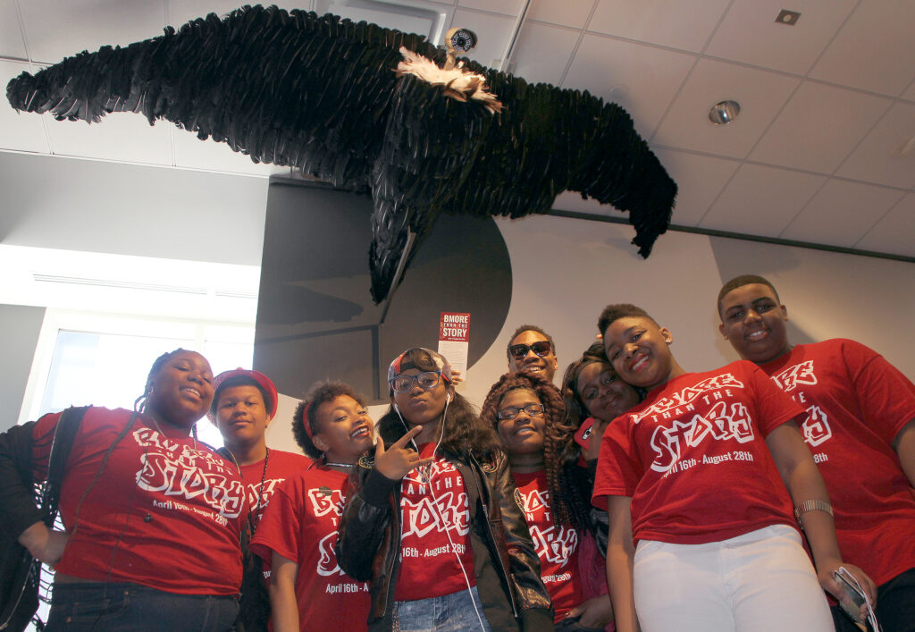 Students in red t-shirts standing below a sculpture of a vulture that has a camera for a head.