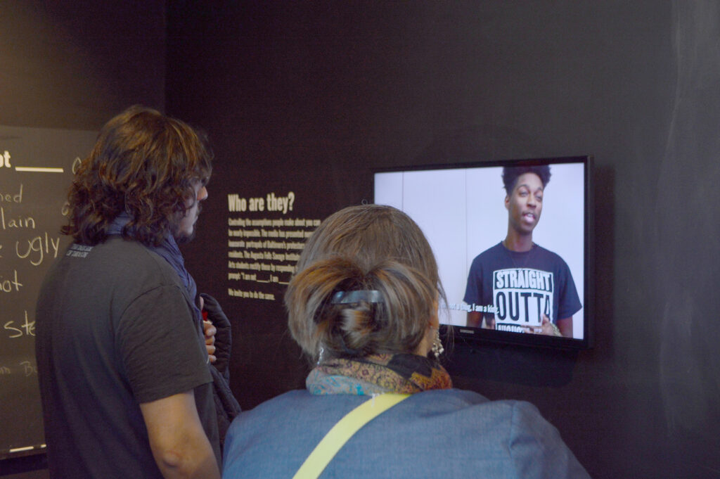 Two people are watching a video of a student speaking in the exhibition.